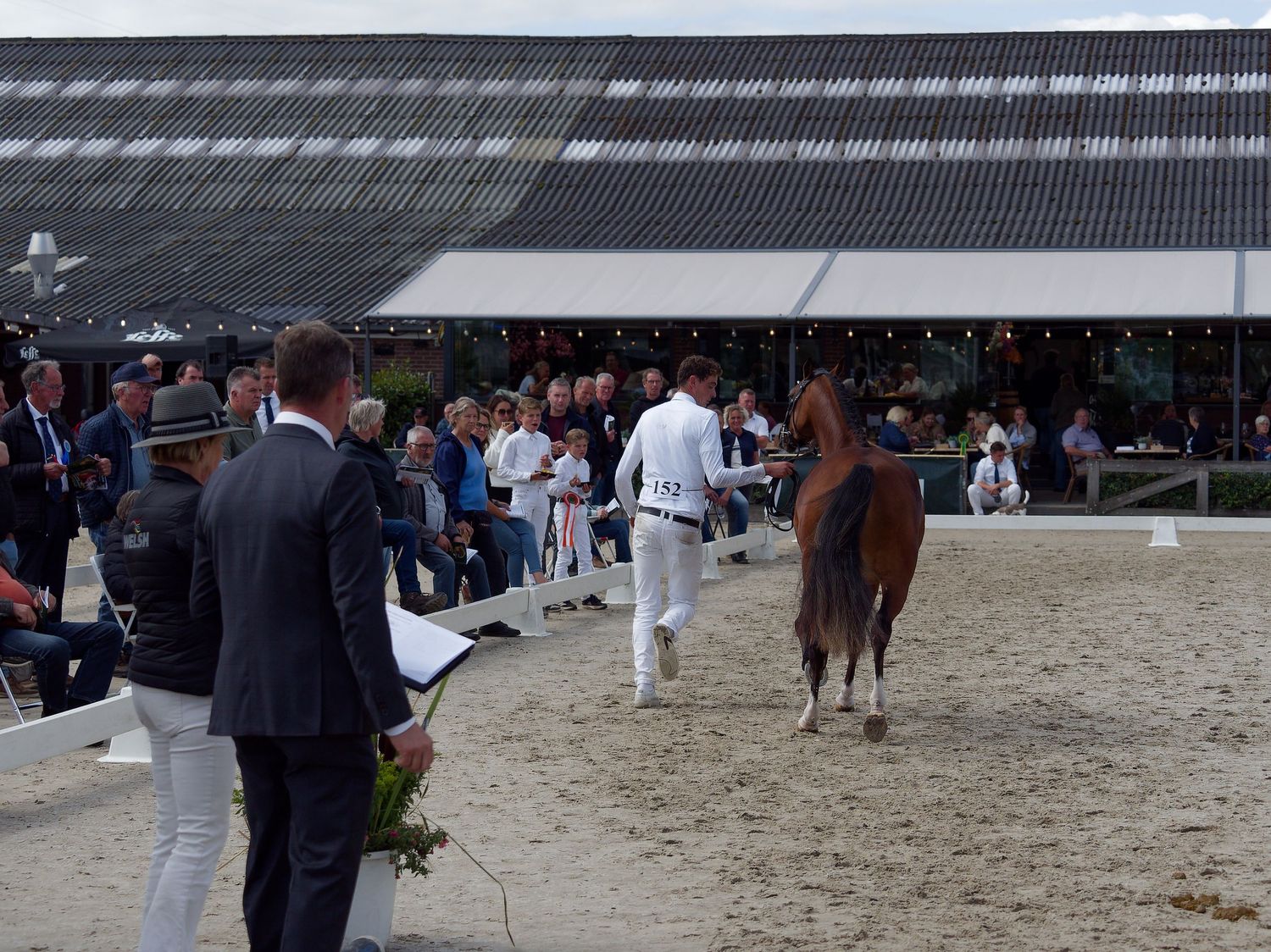 Horse show participants and spectators gather around a sandy arena during competition.