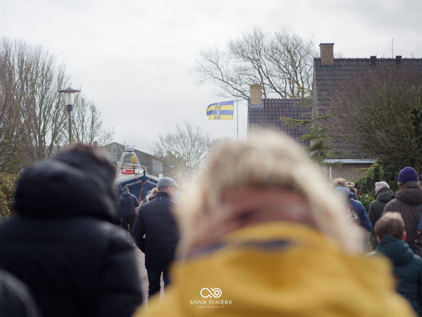 A large crowd gathers in a line outside on an overcast day with buildings and bare trees visible in the background.