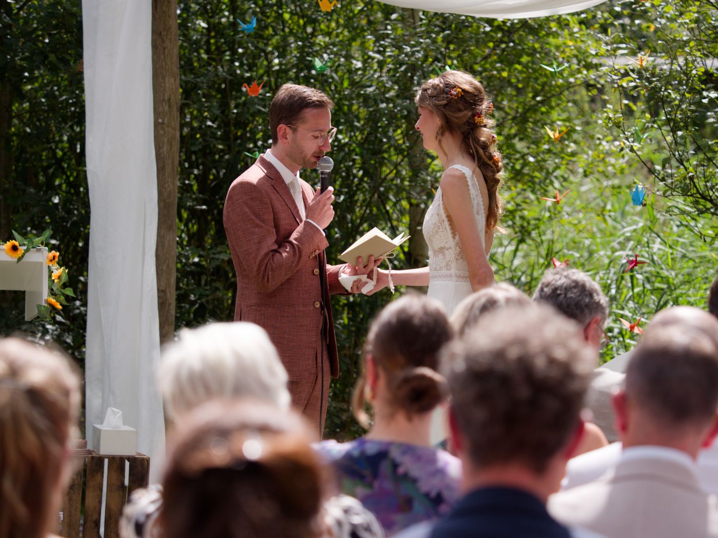 Outdoor wedding ceremony with groom in brown suit reading vows to bride in white dress, surrounded by seated guests.