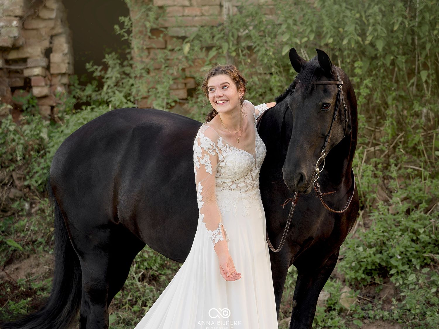 Bride in white lace gown smiling beside a black horse outdoors near rustic stone ruins.