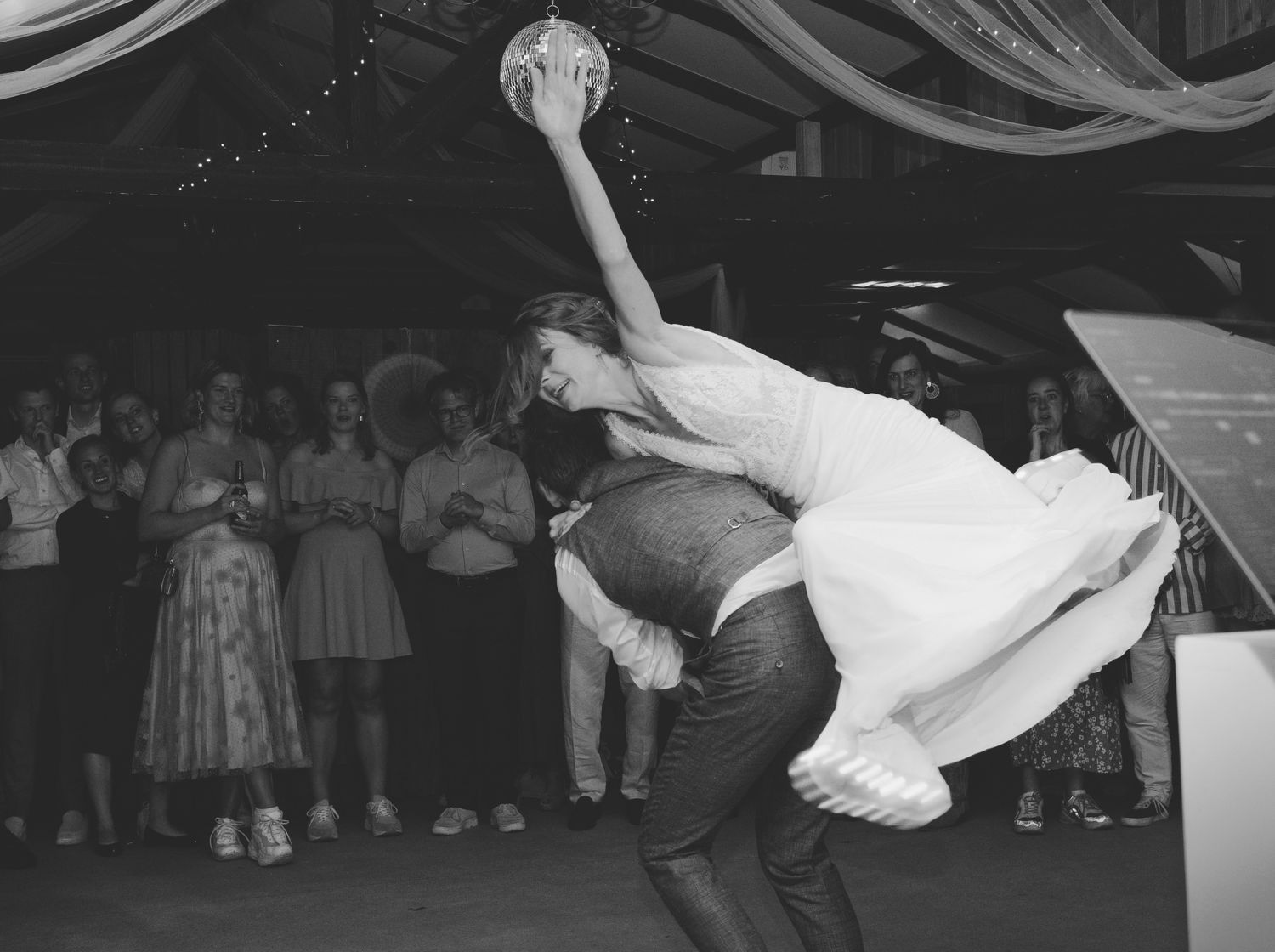 Black and white photo of bride and groom performing dramatic dance lift at wedding reception with guests watching.