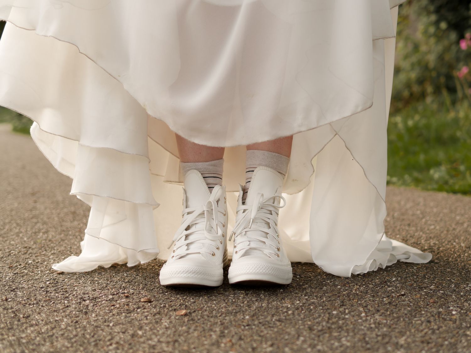 Bride wearing white Converse sneakers under a layered wedding dress on a paved outdoor path.