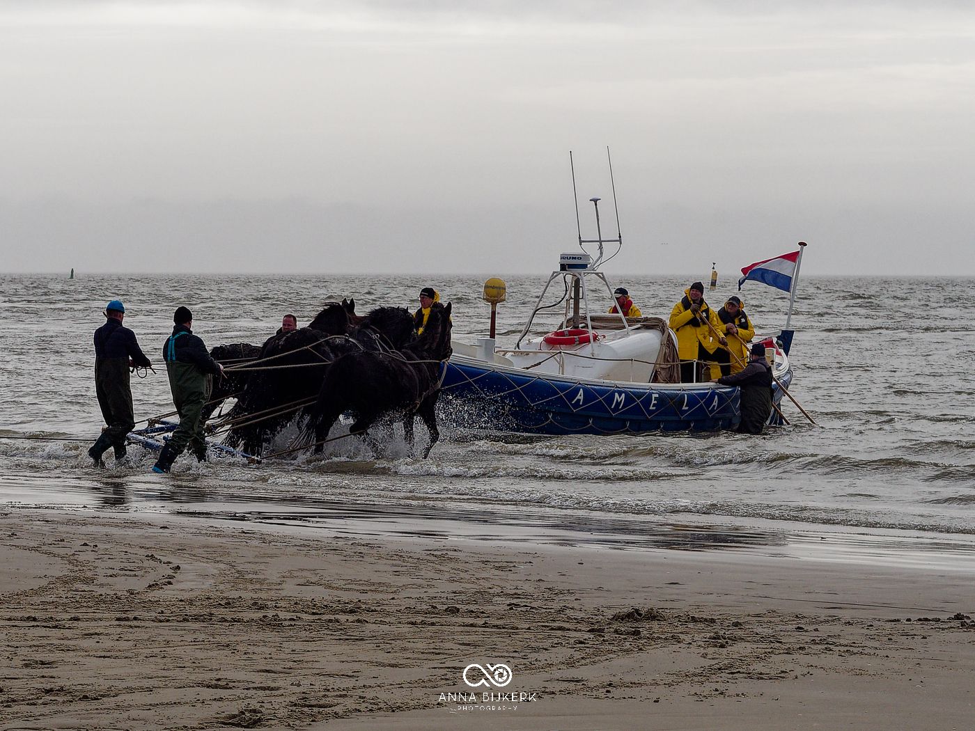 Rescue workers in yellow gear assist with pulling a boat onto a beach during stormy weather conditions.
