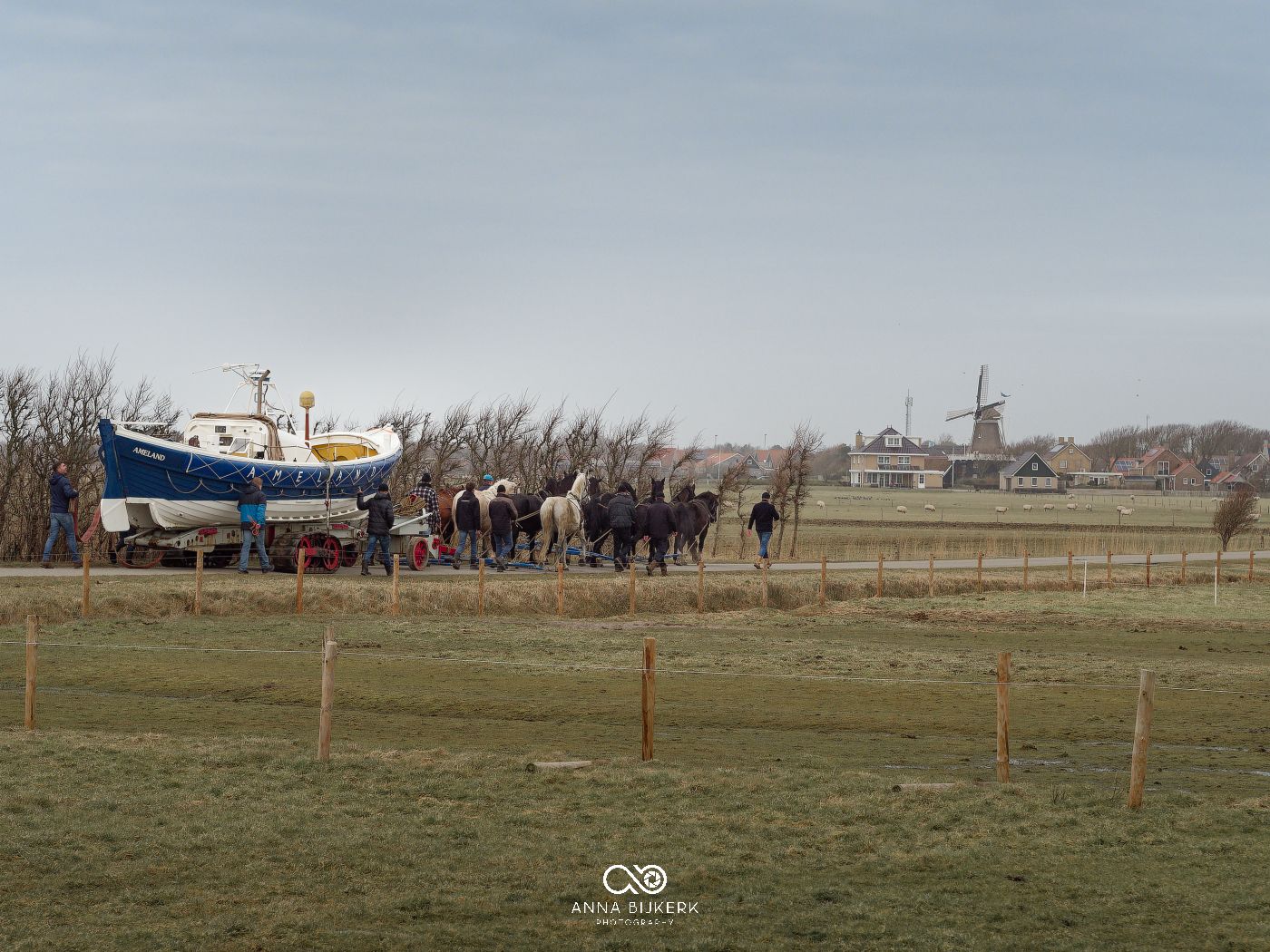 A boat and group of horses stand in a grassy field with a windmill visible in the distant landscape.