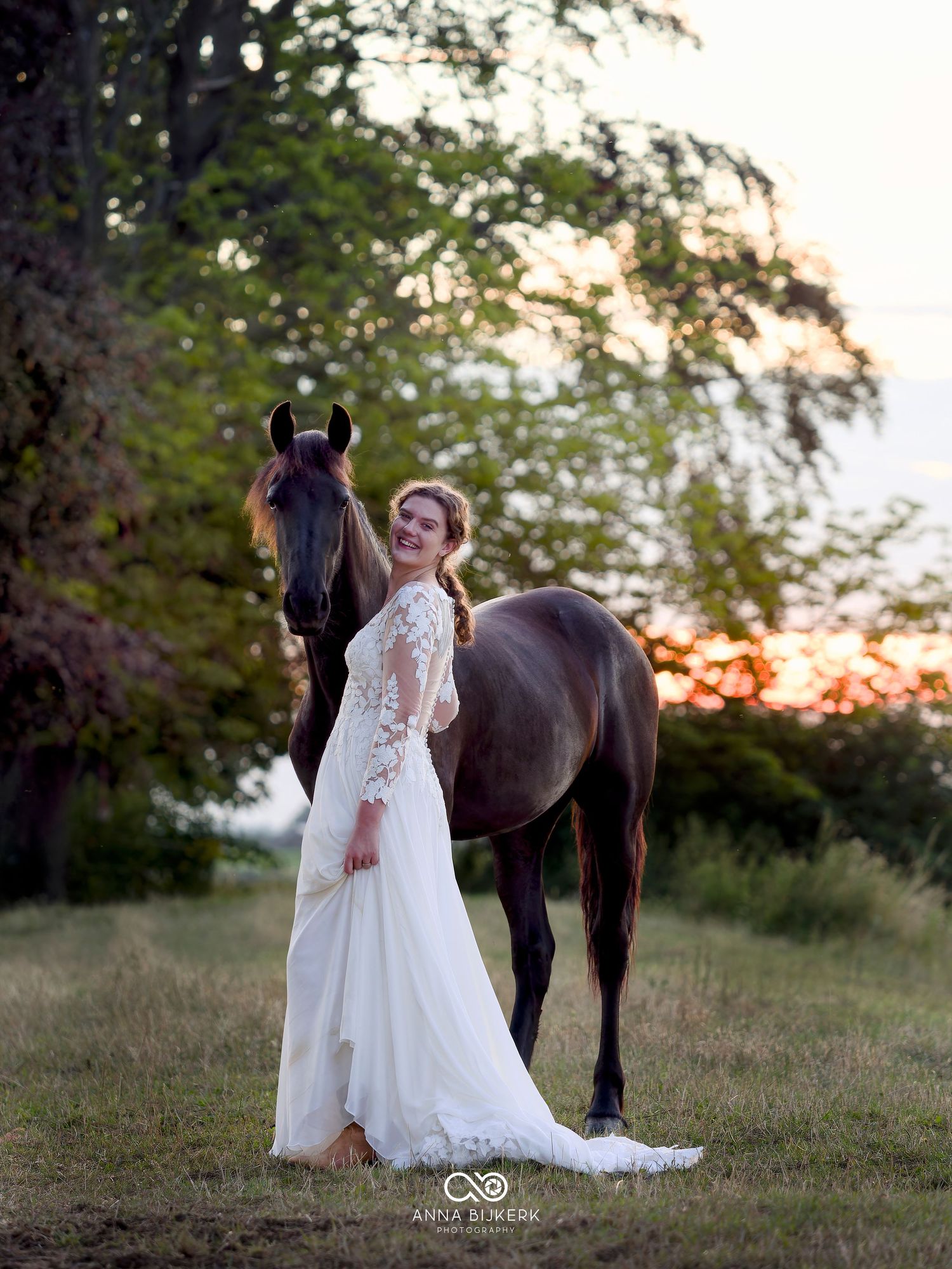 Bride in white lace gown stands beside a dark horse at golden hour in a lush green field.