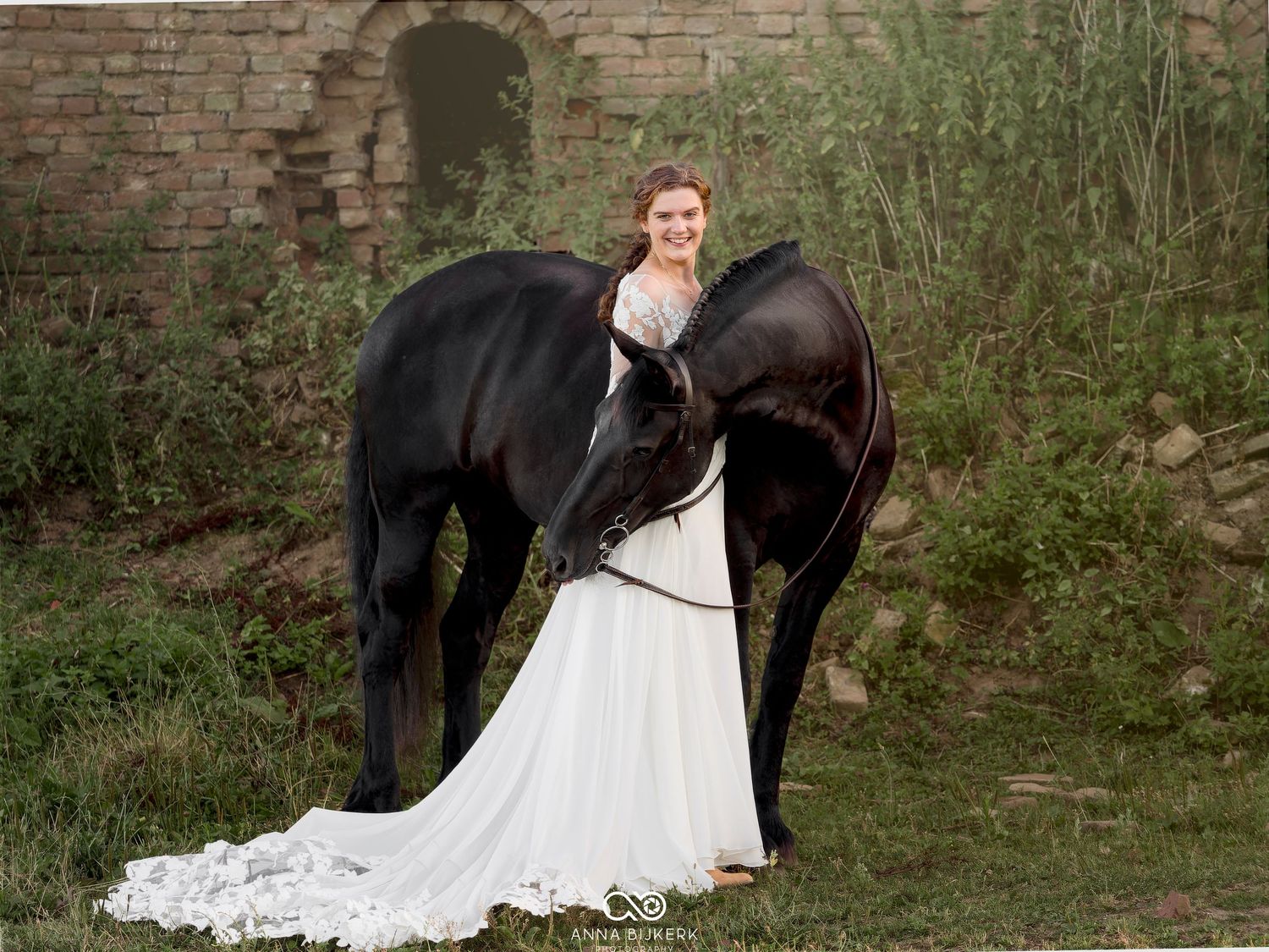 Bride in white gown poses with black horse near stone ruins surrounded by lush greenery.