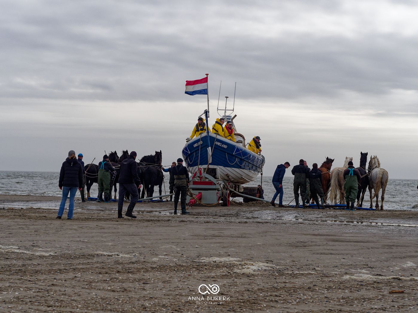 A group of people gathered around a fishing boat on a cloudy beach with a Dutch flag flying overhead.