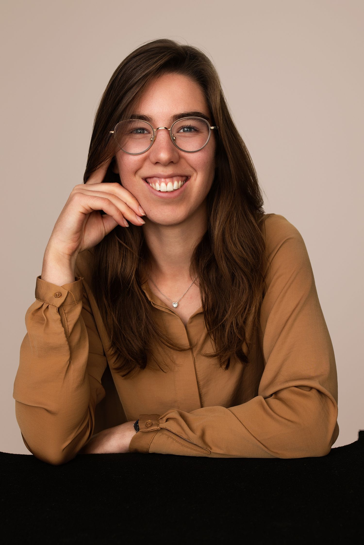 Smiling young woman with glasses and brown hair wearing a tan blouse, posing with hand on chin against beige background.