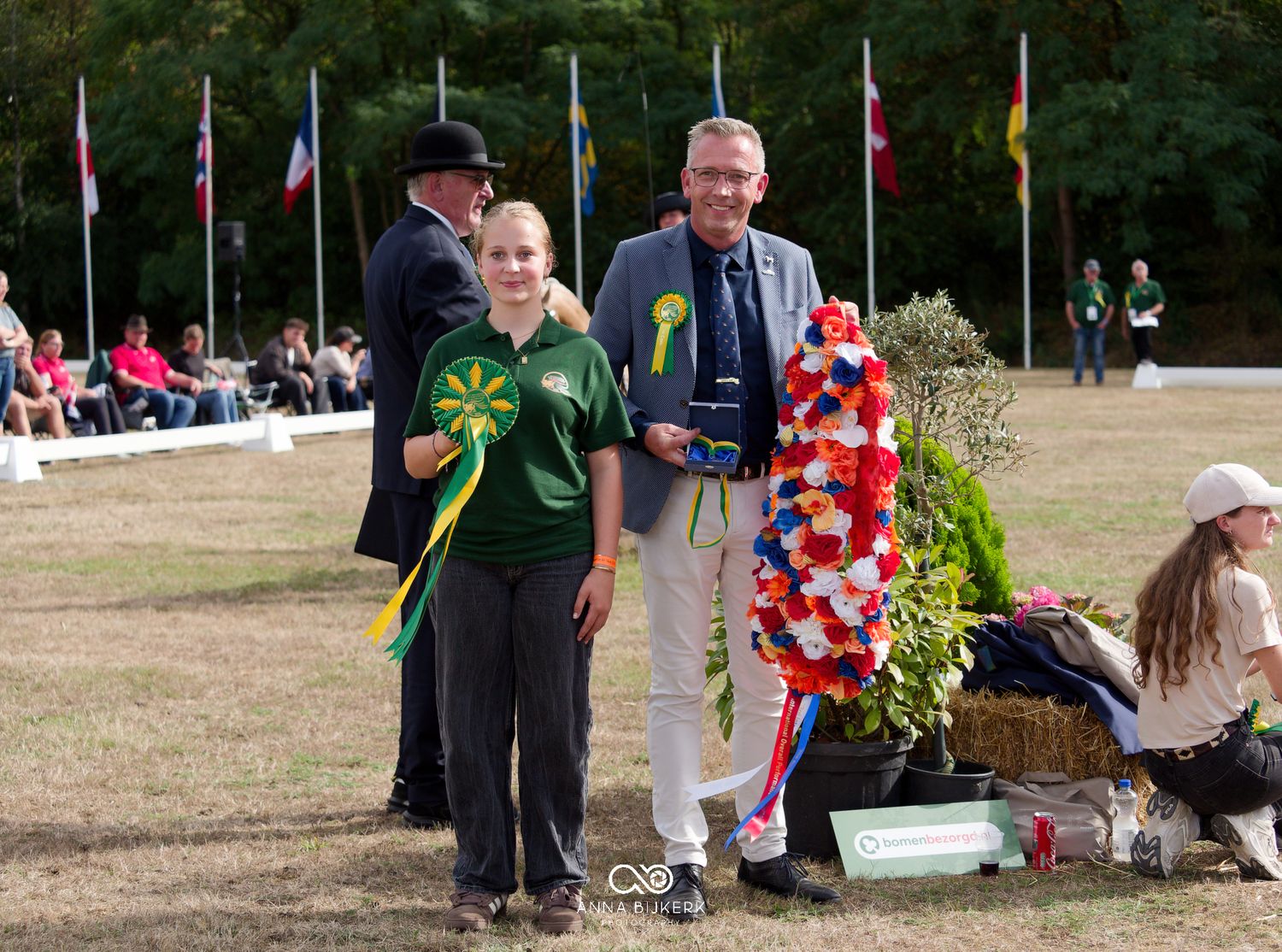Two award winners pose with colorful rosettes at an outdoor equestrian competition surrounded by international flags.