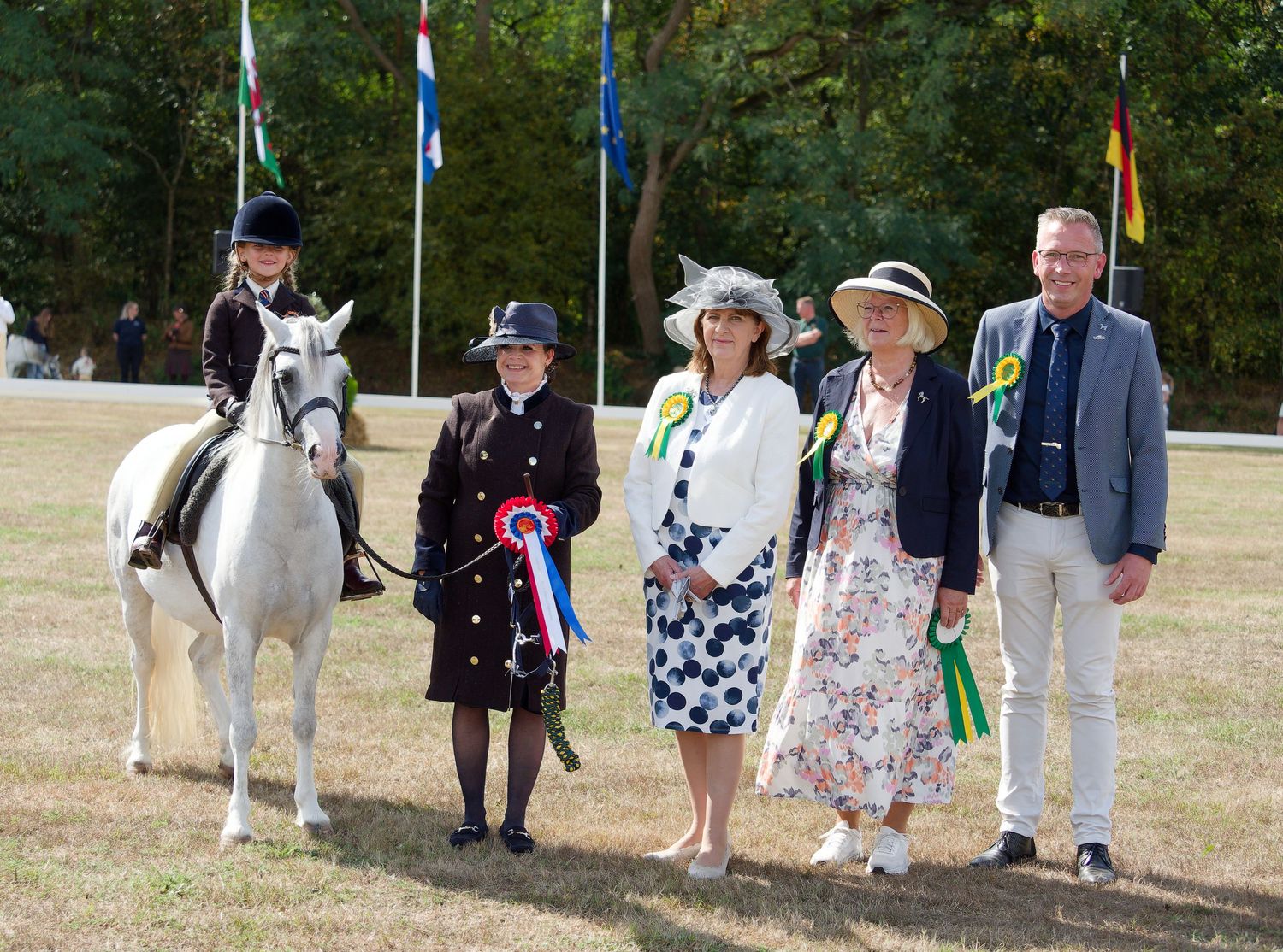 Young equestrian on white horse poses with judges and officials at an outdoor horse show competition.