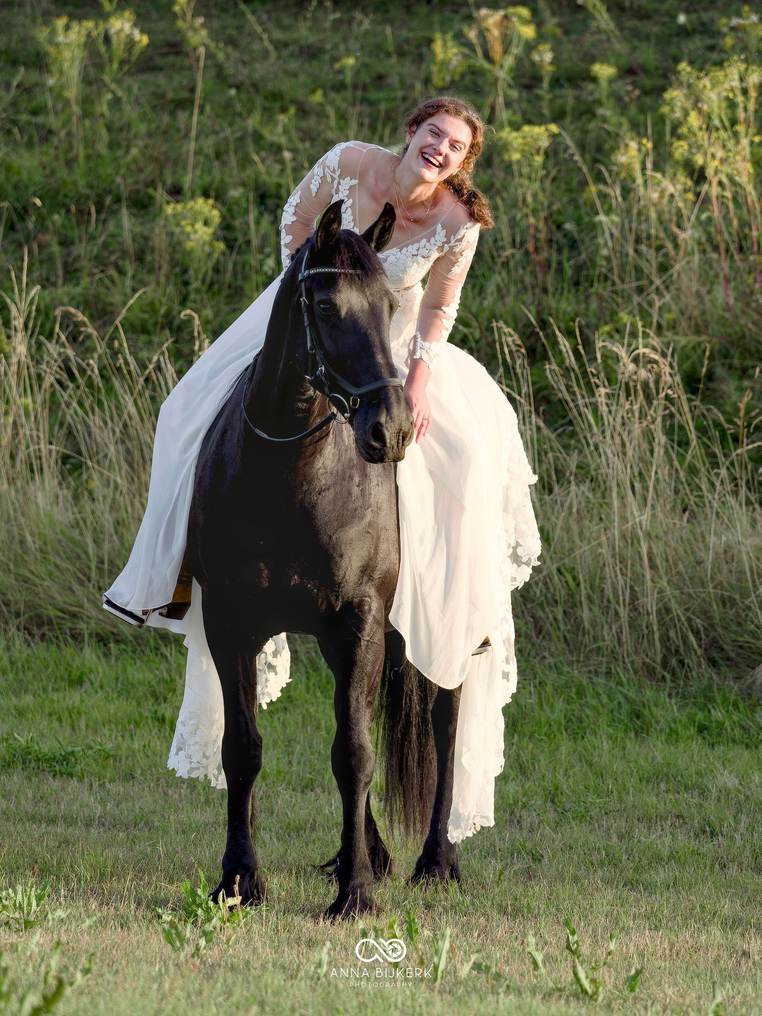 A laughing bride in a white lace gown rides a black horse through a green meadow.