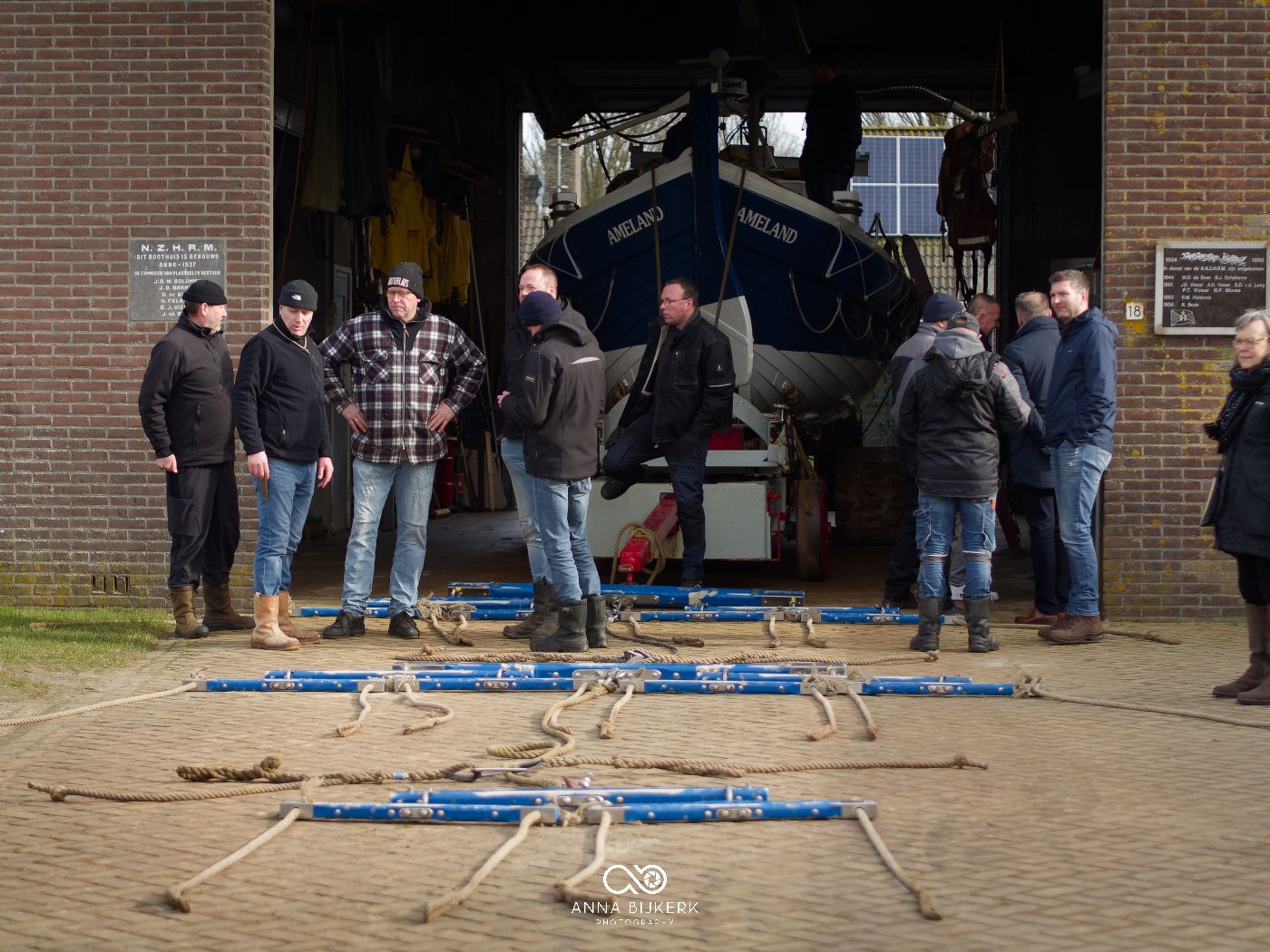 A group of people gather around a boat on stands at a boatyard with maintenance equipment laid out on the ground.