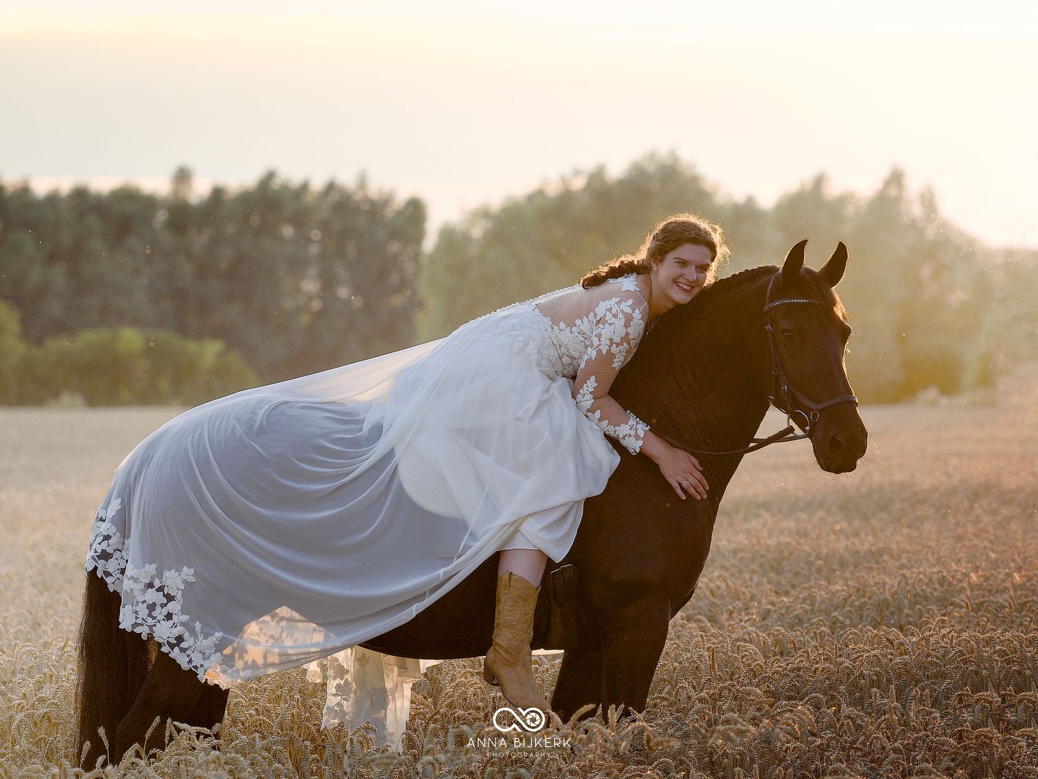 Bride in white wedding gown and veil leans over dark horse in golden wheat field at sunset.