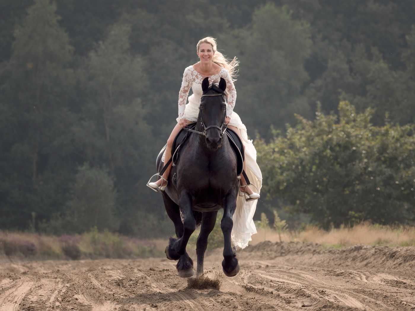 Blonde woman in white wedding dress riding a dark horse galloping along a dirt path with trees in background.