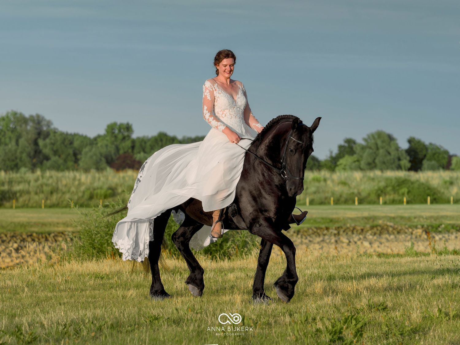 Bride in white lace gown riding a black Friesian horse across a green meadow on a sunny day.