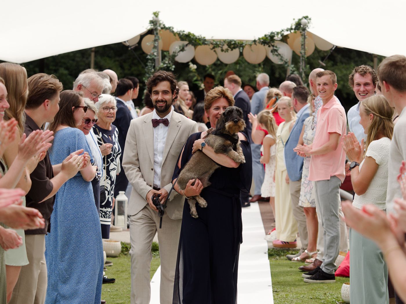 Couple walks down wedding aisle holding dog, guests clapping, outdoor ceremony with floral arch backdrop.