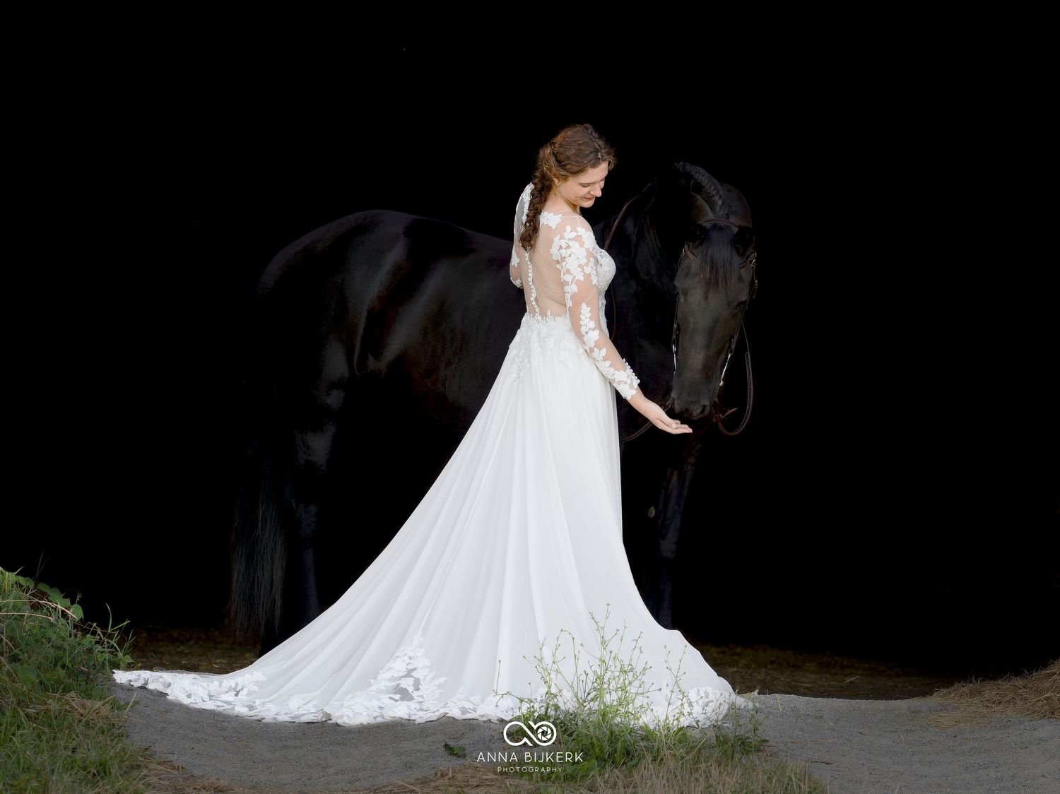 Bride in white lace gown with long train stands beside a dark horse against a black background.