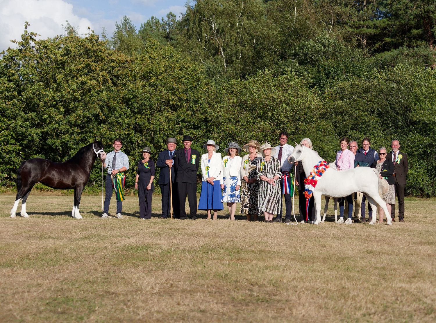 Group of people posing with two horses, a dark and white one, at an outdoor equestrian show on a sunny day.