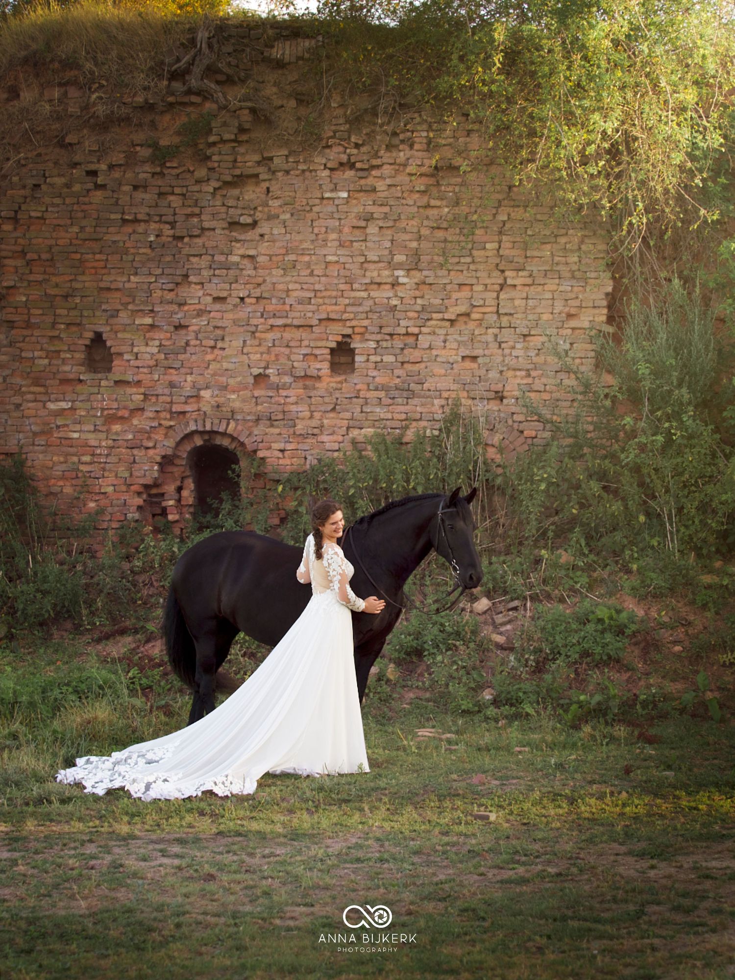 Bride in white gown with long train stands beside a black horse near rustic brick ruins in a lush garden setting.