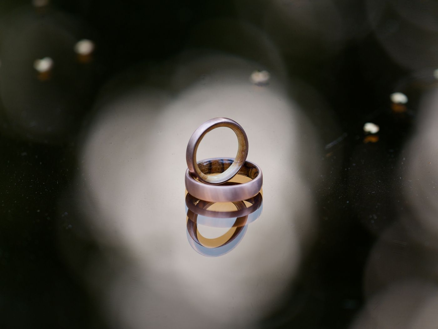 Two gold wedding bands stacked and reflected on a dark surface with bokeh background lighting.