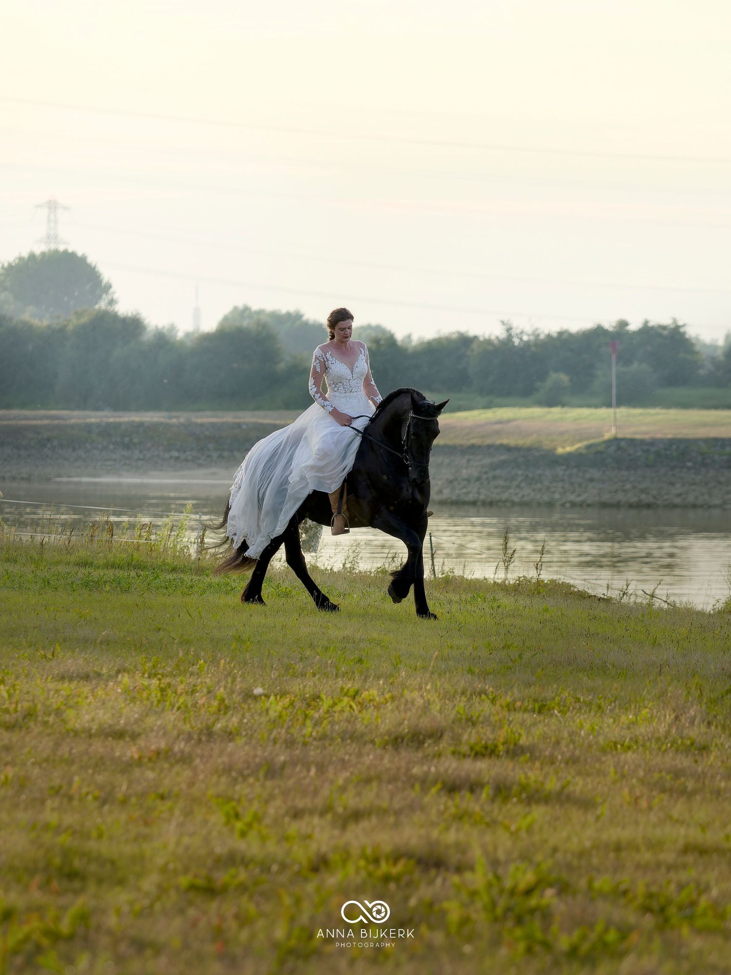 Bride in white wedding dress riding a black horse along a riverbank at golden hour.
