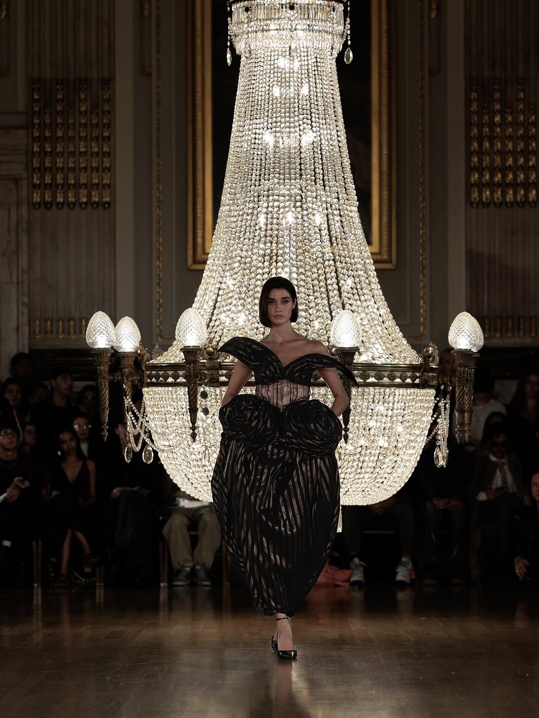 A figure performs beneath an elegant crystal chandelier in a grand ballroom setting during what appears to be a performance.