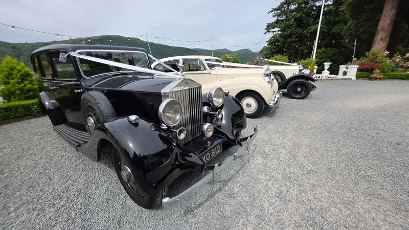 Vintage classic black and cream Rolls-Royce wedding cars parked on a driveway with green hills in the background.