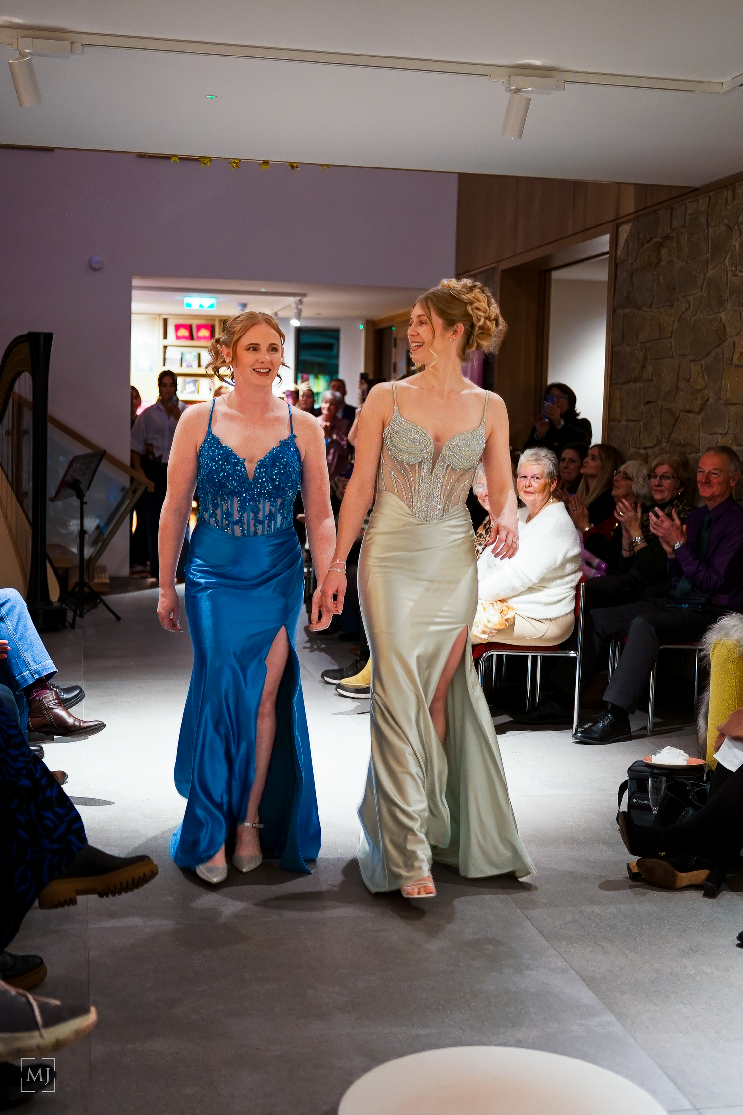 Two people in elegant evening gowns, one blue and one silver, walking together in a reception hall with guests watching.