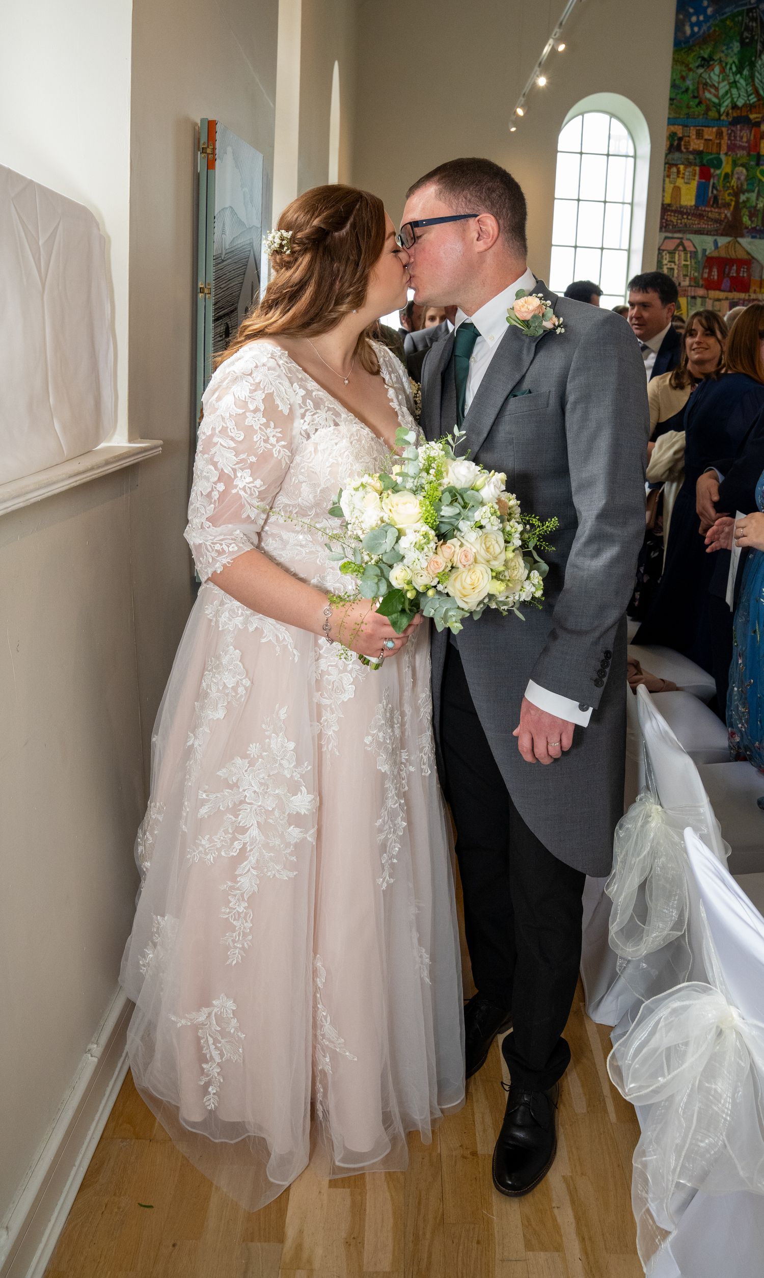 Bride and groom share a kiss at their wedding ceremony, bride holding a white floral bouquet in a lace gown.