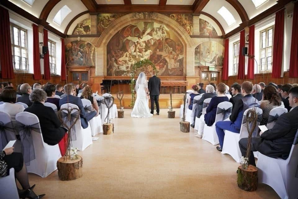 Elegant wedding ceremony in a grand hall with ornate murals, white chair covers, and guests seated along a central aisle.