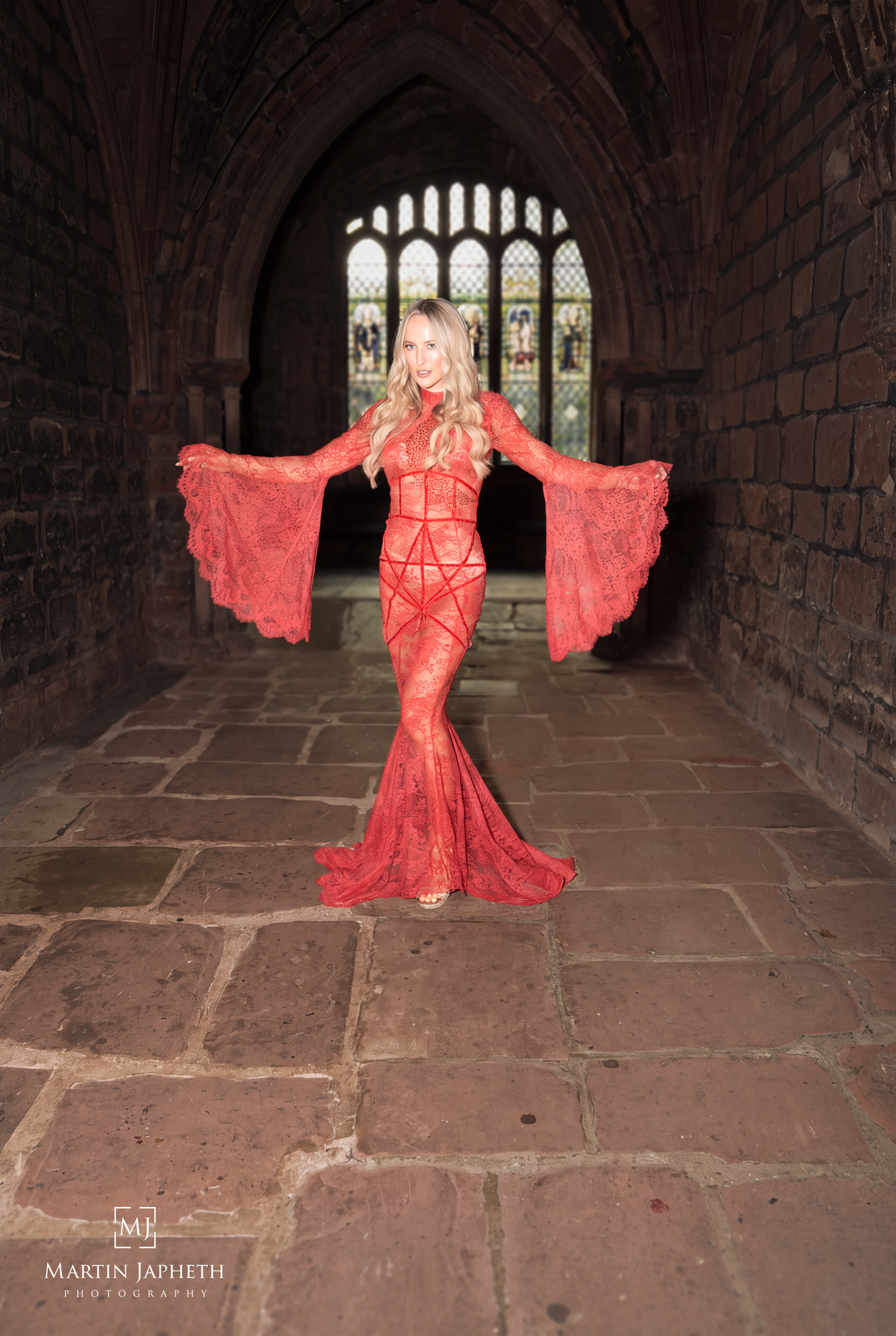 Woman in red lace mermaid gown with dramatic sleeves poses in stone archway with stained glass window.
