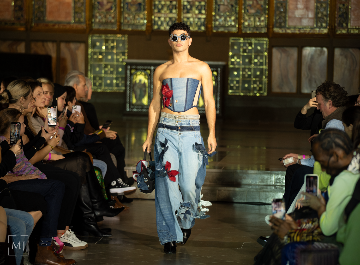 A model walks the runway wearing a denim and corset ensemble during a fashion show with audience members seated along sides.