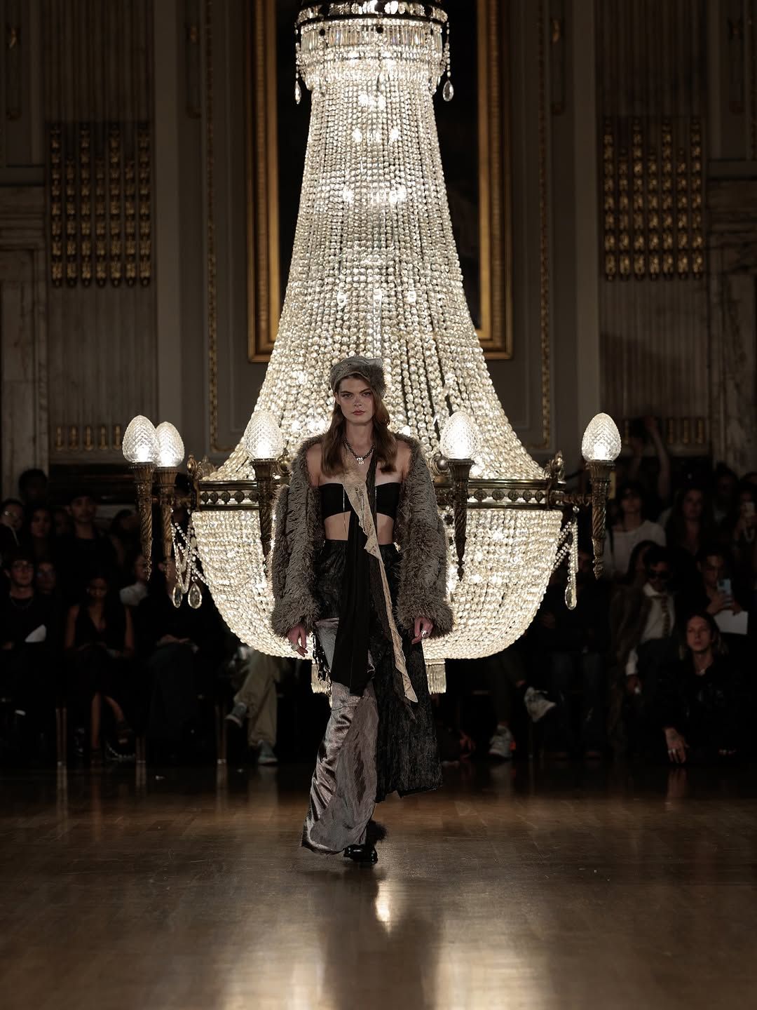 A model walks beneath a grand crystal chandelier during a dramatic fashion show presentation.