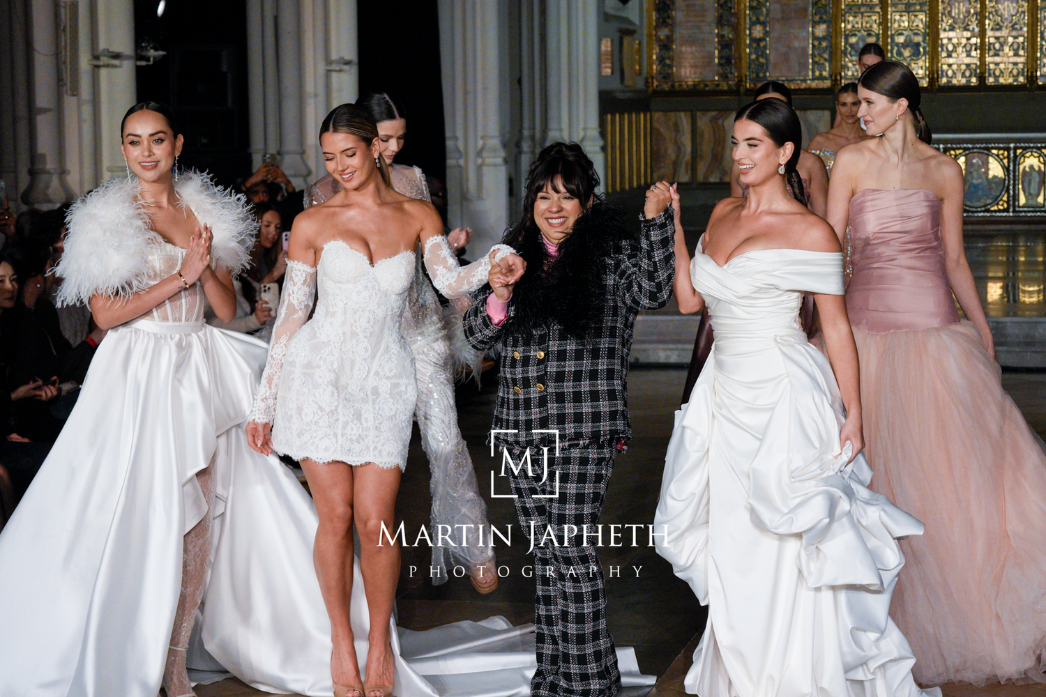 Models walk the runway showcasing elegant white wedding gowns and blush bridesmaid dresses during a bridal fashion show.
