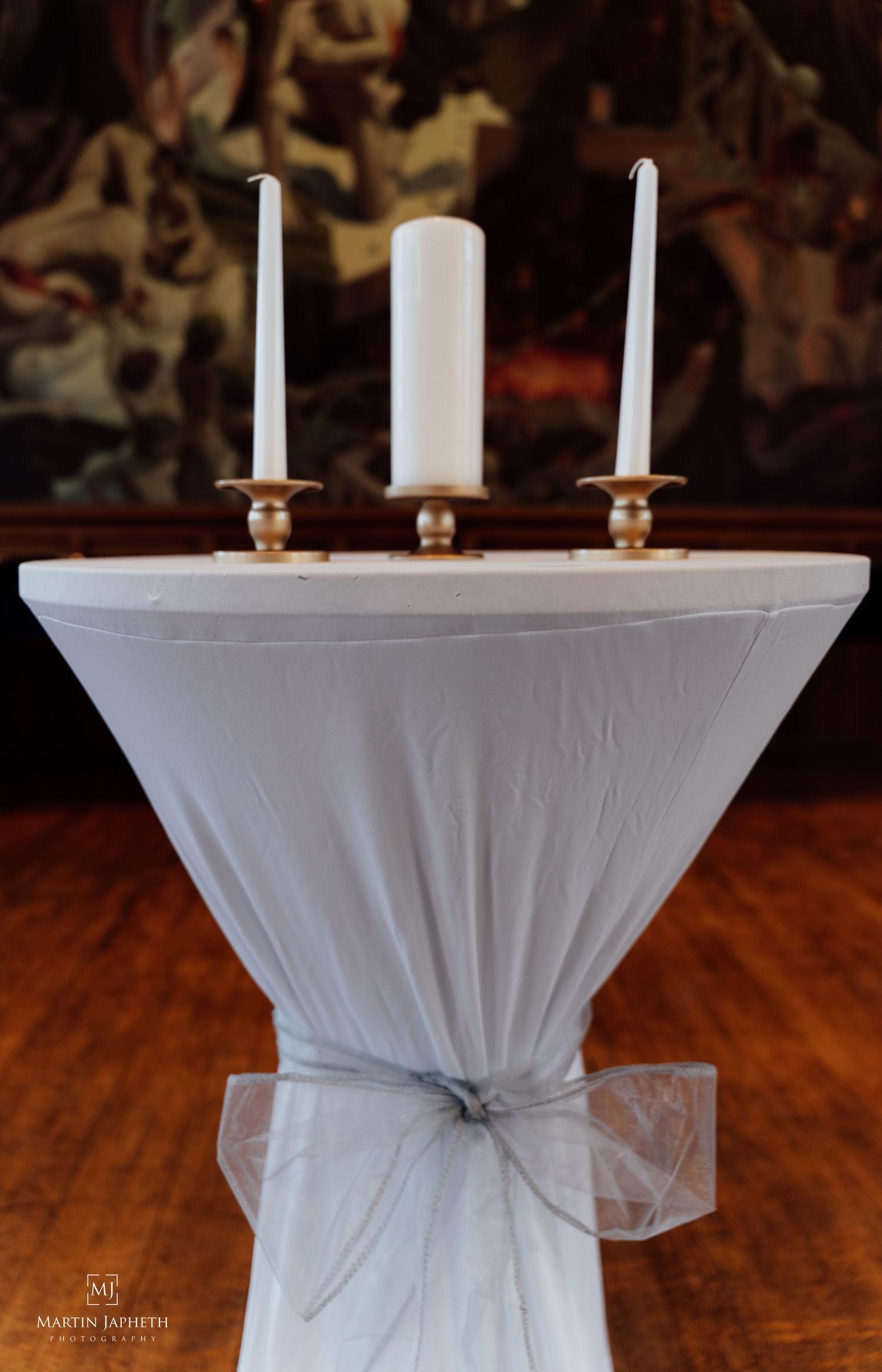 Elegant unity candle set on a draped white cocktail table with tulle bow, set against a decorative painting backdrop.
