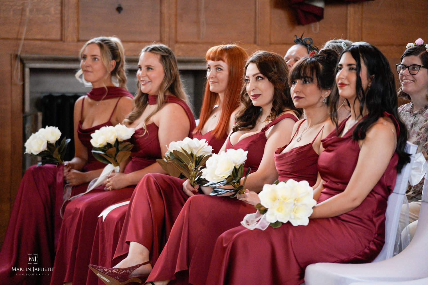 Six bridesmaids in matching red dresses holding white bouquets seated at a wedding ceremony. Martin Japheth Photography