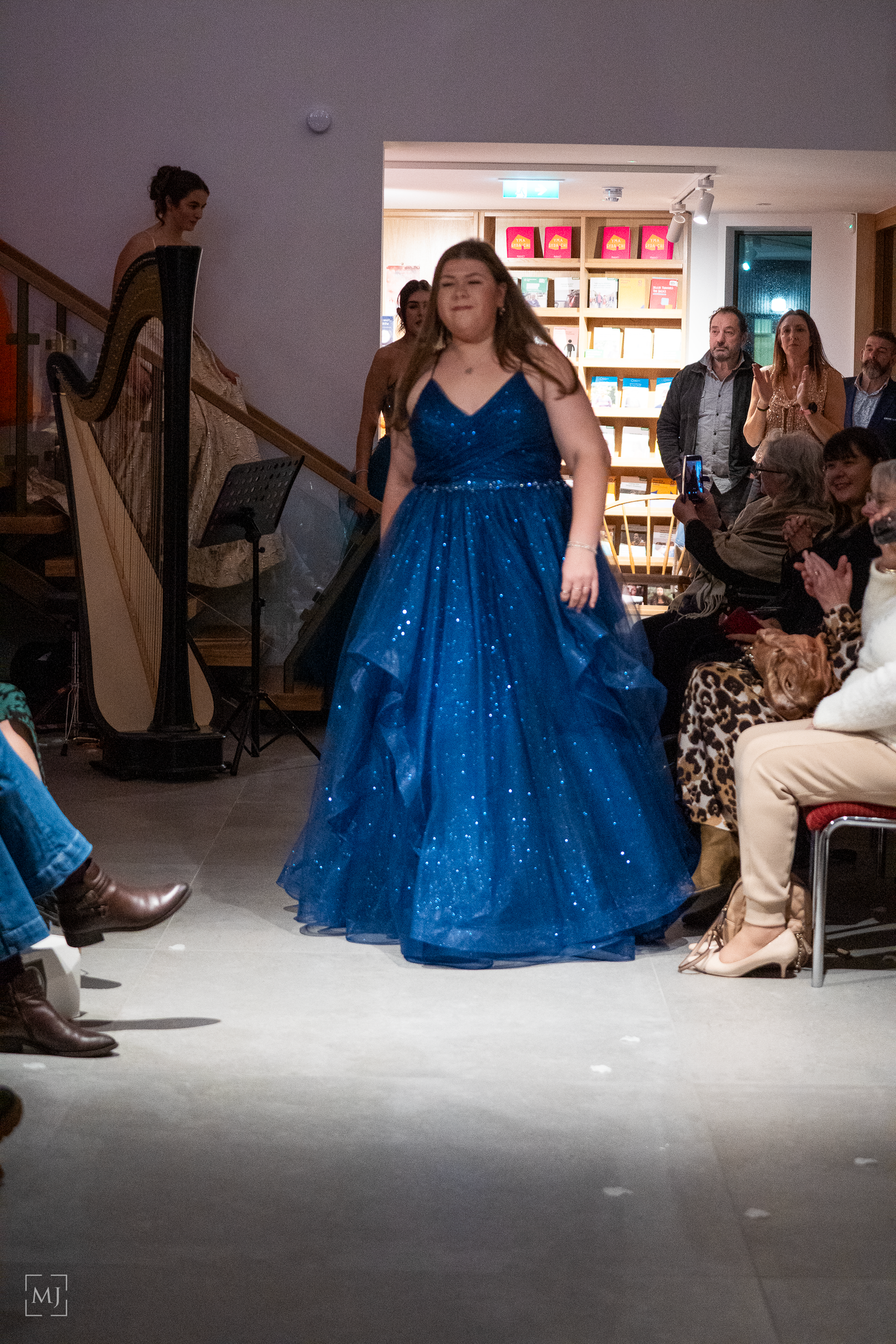 A model walks the runway in a sparkling royal blue ball gown at a fashion show while a harpist plays in the background. Fashion Photographer