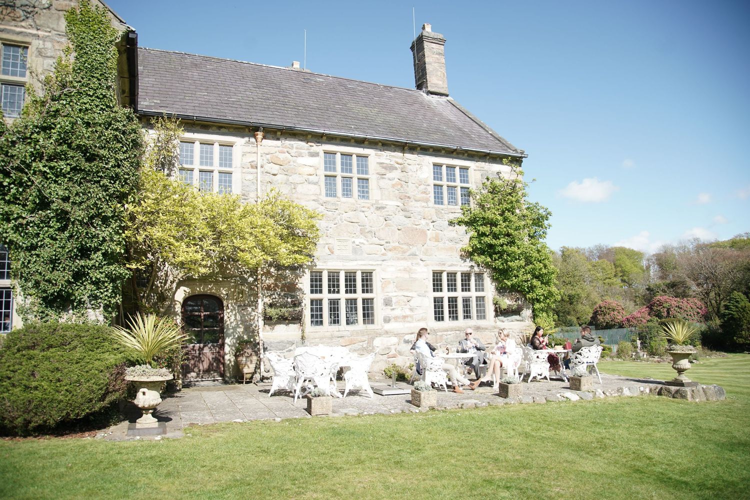 Stone manor house with guests relaxing on sunny patio surrounded by lush green lawn and mature trees on a clear day.