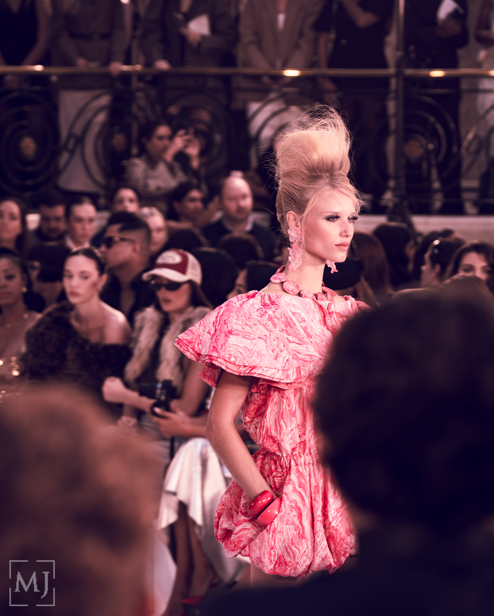 Model walks runway in pink ruffled dress during high fashion show with crowd in background.