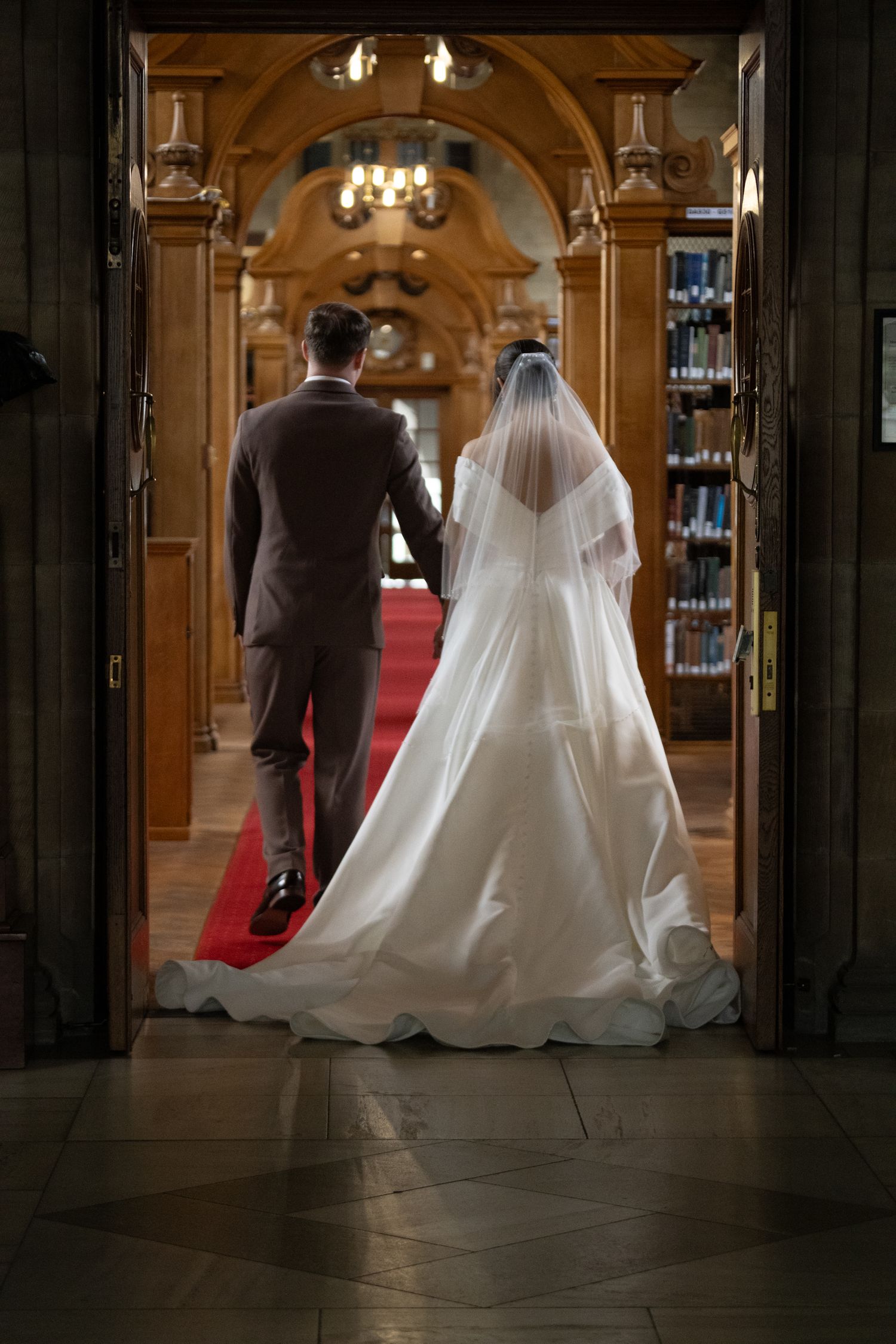 Bride and groom walking hand in hand down a grand ornate hall with red carpet and arched ceilings.