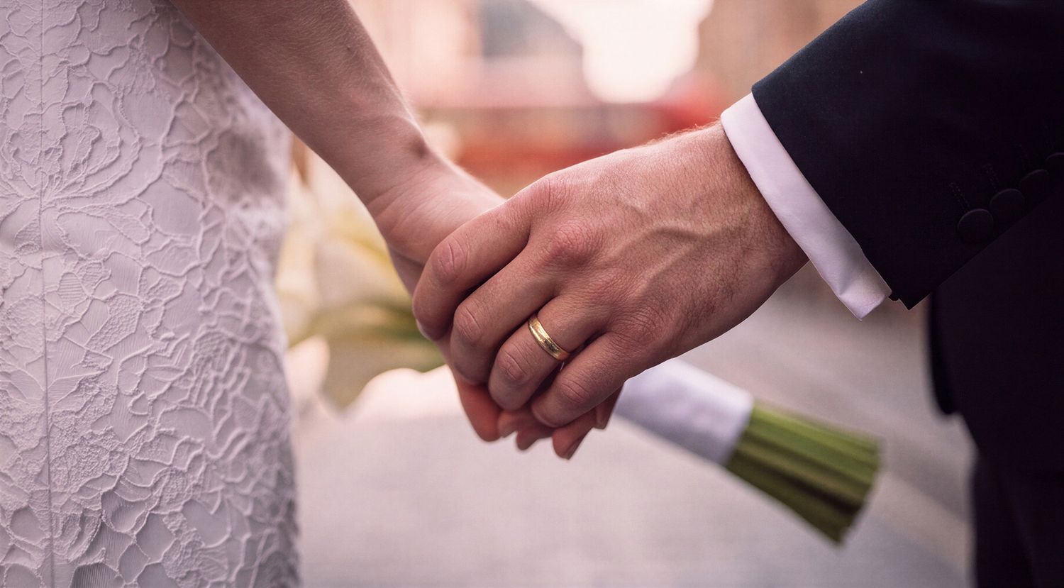 Bride and groom holding hands at wedding ceremony, bride in lace dress, groom in suit with gold wedding ring.