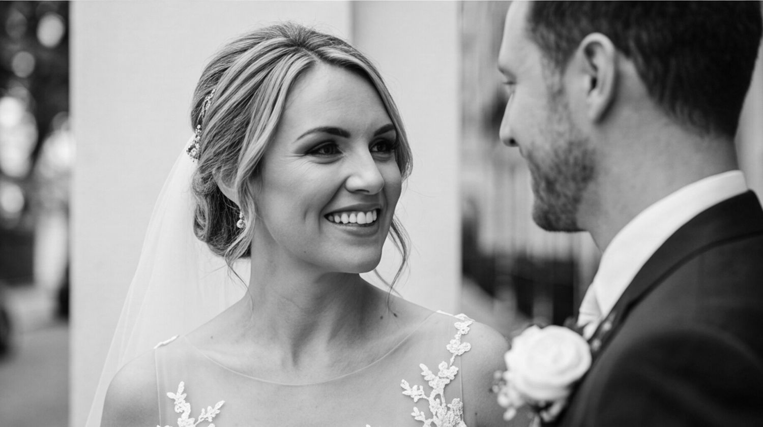 Black and white wedding photo of a smiling bride in lace gown gazing at groom in suit with boutonniere.