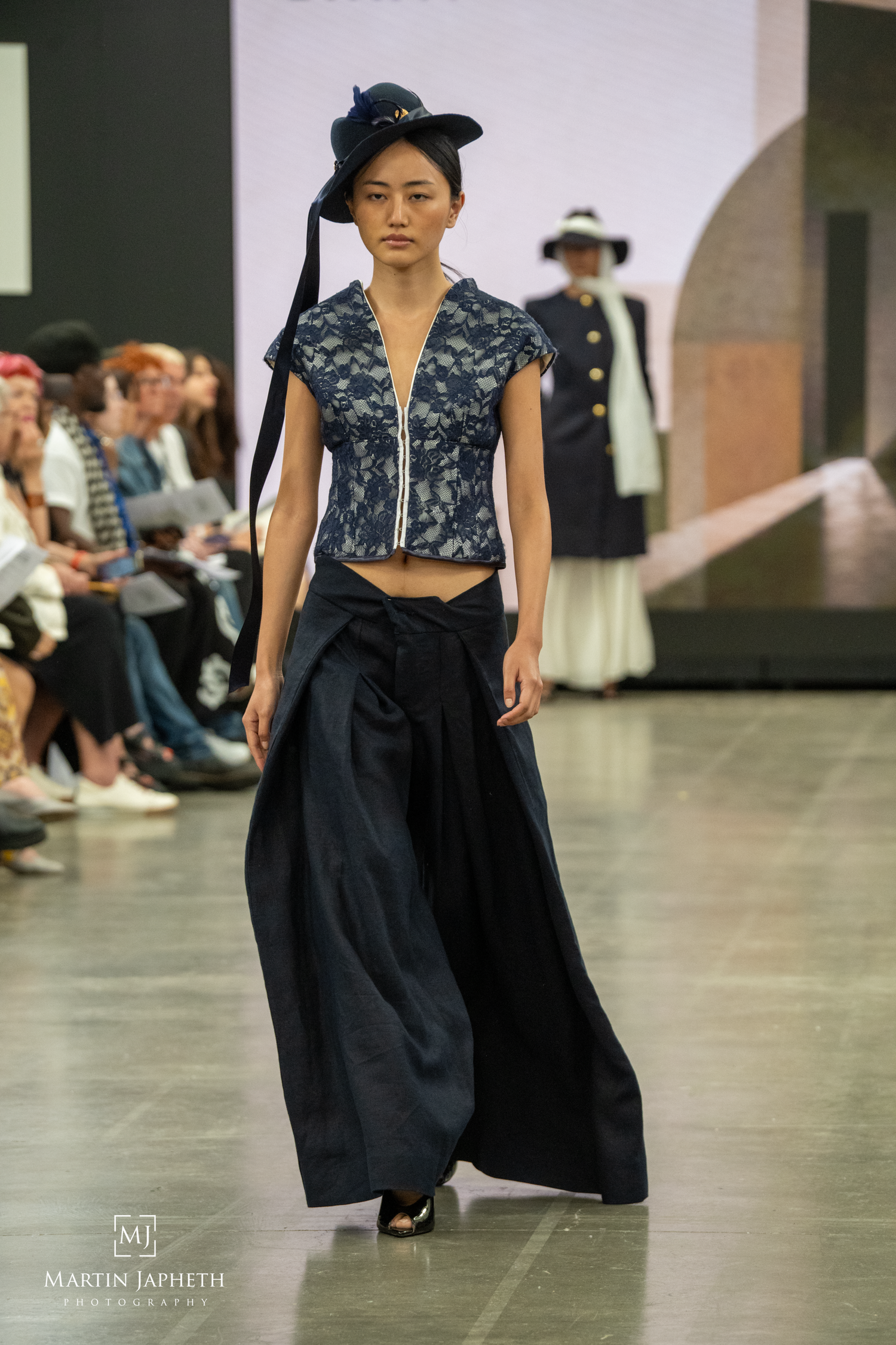 Model walks runway in navy lace crop top, wide-leg trousers, and tilted hat at a fashion show. Martin Japheth Photography