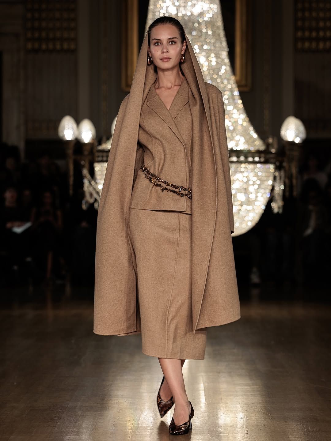 Fashion model walks runway in camel-colored wrap coat and dress ensemble with matching headpiece at luxury show.