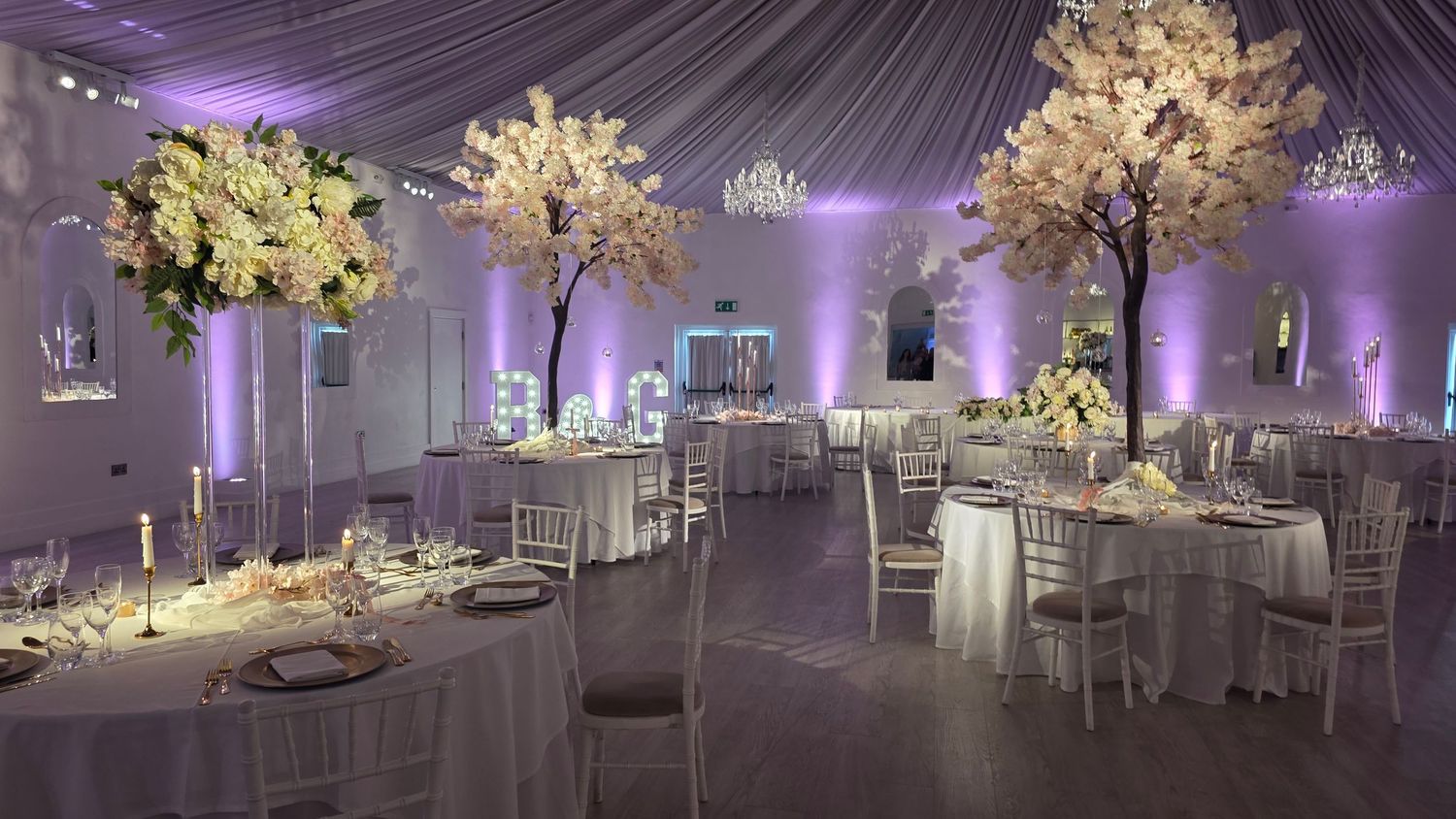 Elegant wedding reception hall with white floral trees, round tables, silver chairs, and soft purple uplighting.