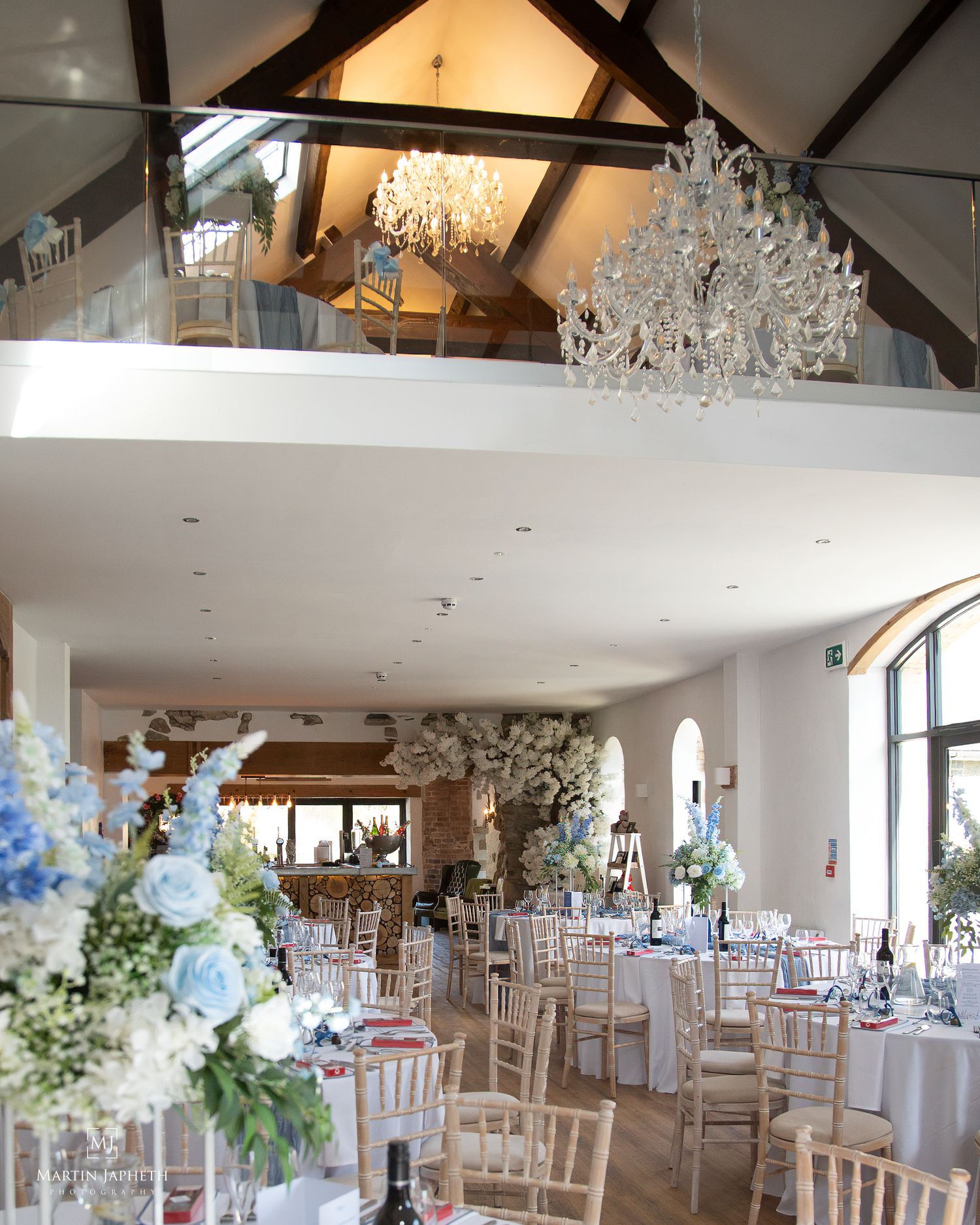 Elegant wedding venue with crystal chandeliers, vaulted beamed ceiling, mezzanine balcony and tables dressed with blue and white floral cent
