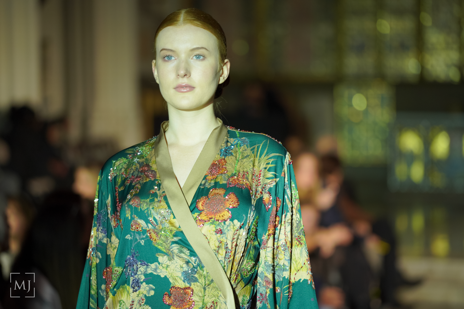 Model walks runway in vibrant teal floral print dress during fashion show presentation.