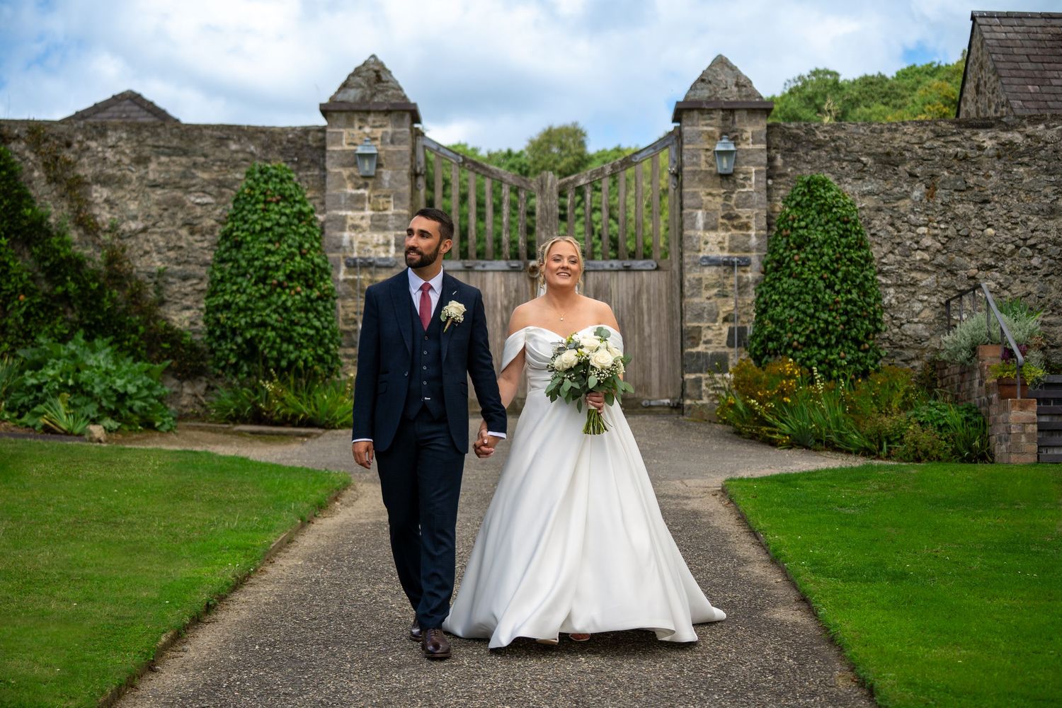 A bride and groom walk hand in hand through a formal garden pathway with stone gates and topiary in the background.
