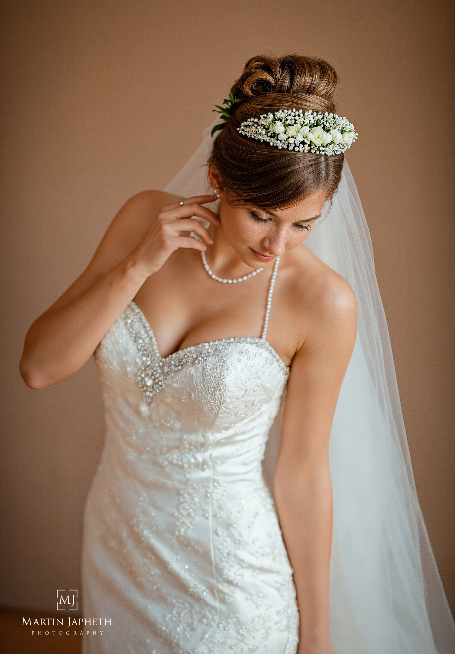 Bride in lace strapless gown with floral crown, veil, and pearl necklace adjusting earring on wedding day.