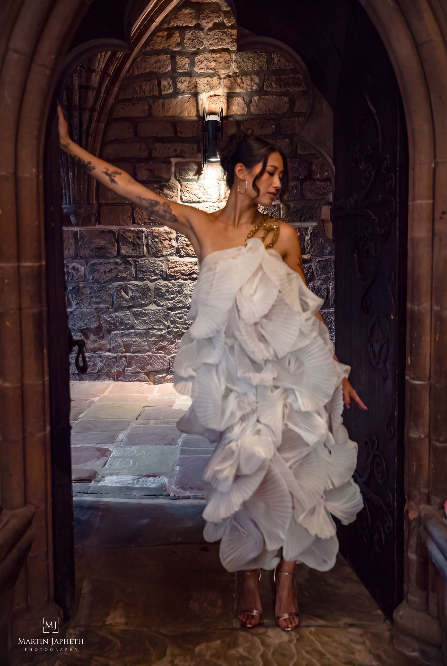 Woman in ruffled white gown poses dramatically in stone archway doorway with warm lighting.